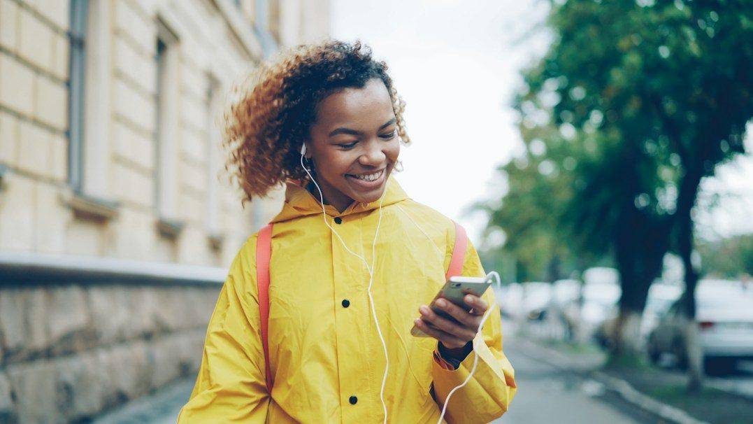 A smiling woman in a yellow raincoat listens with earphones while looking at her smartphone