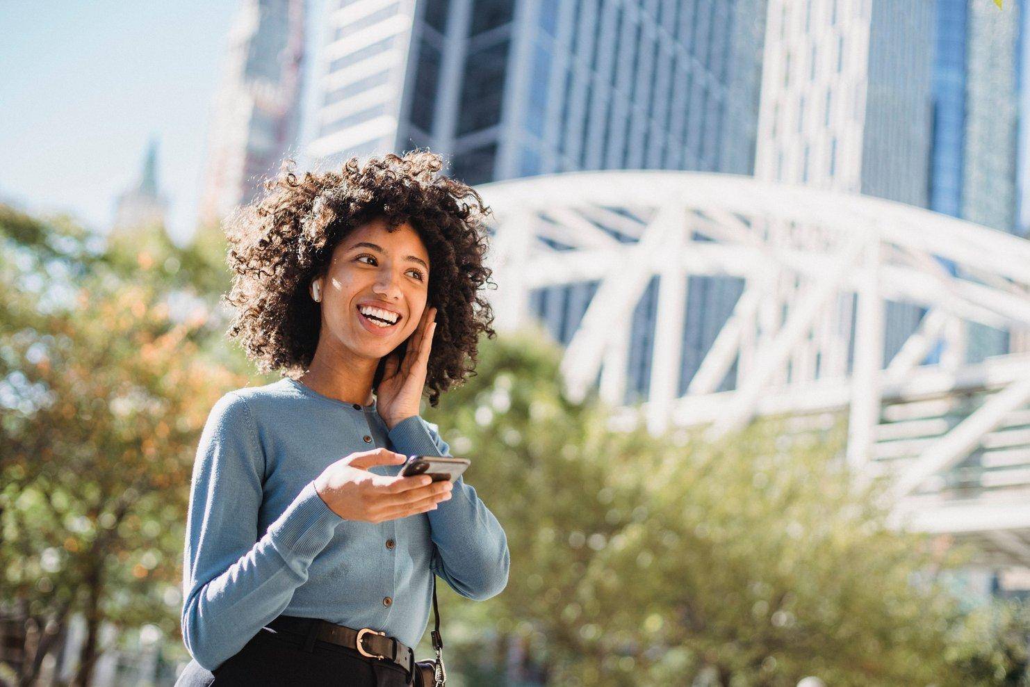 A smiling woman with earphones holds her phone on a city bridge