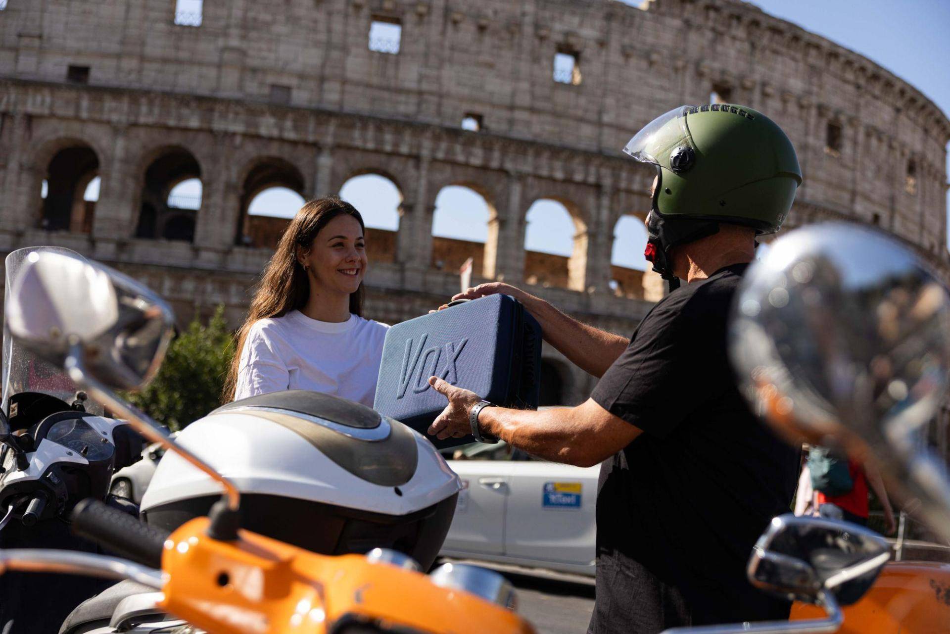 A courier on a scooter hands a Vox equipment case to a tour guide in front of the Colosseum